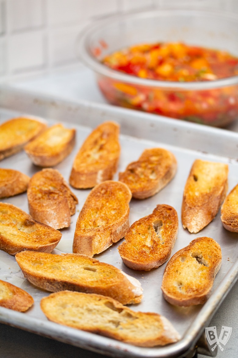 Tray of toasted baguette with bowl of tomato bruschetta mixture in the background