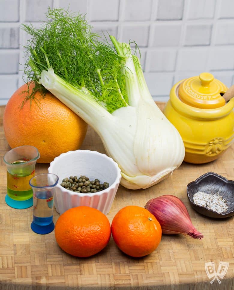 Ingredients for fennel and citrus salad with shallots and capers out on a cutting board.