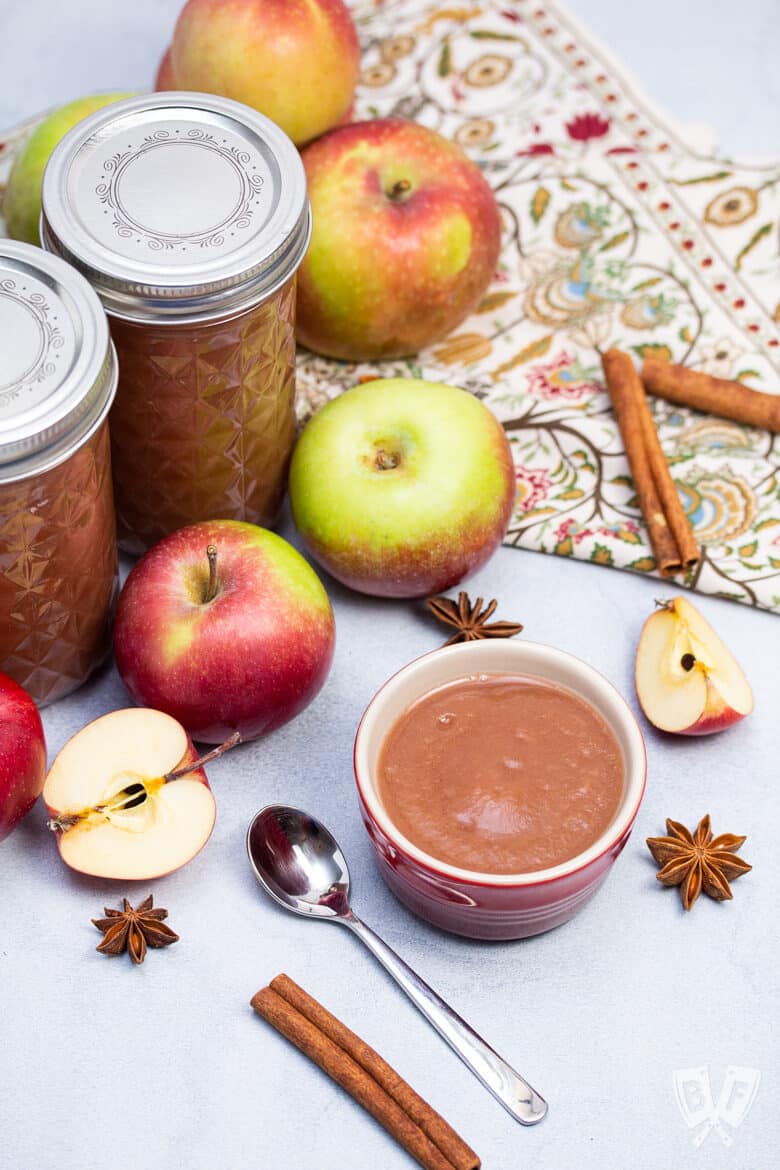 A small bowl of homemade apple sauce with mulling spices, apples, and jars of applesauce.