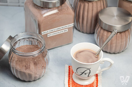 Homemade hot cocoa in a mug surrounded by jars of hot cocoa mix.