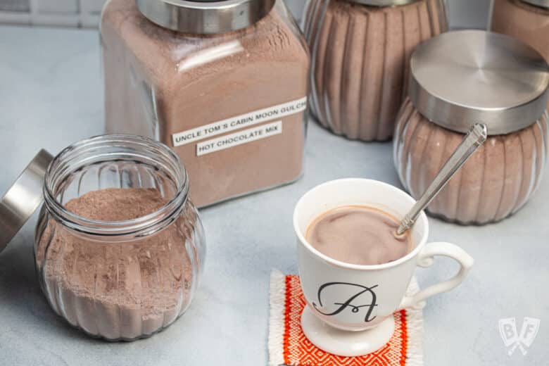 Homemade hot cocoa in a mug surrounded by jars of hot cocoa mix.