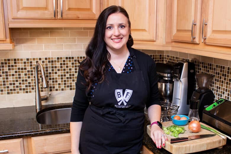 Ashley Covelli standing in the kitchen next to a cutting board full of ingredients, wearing an apron with the Big Flavors logo.