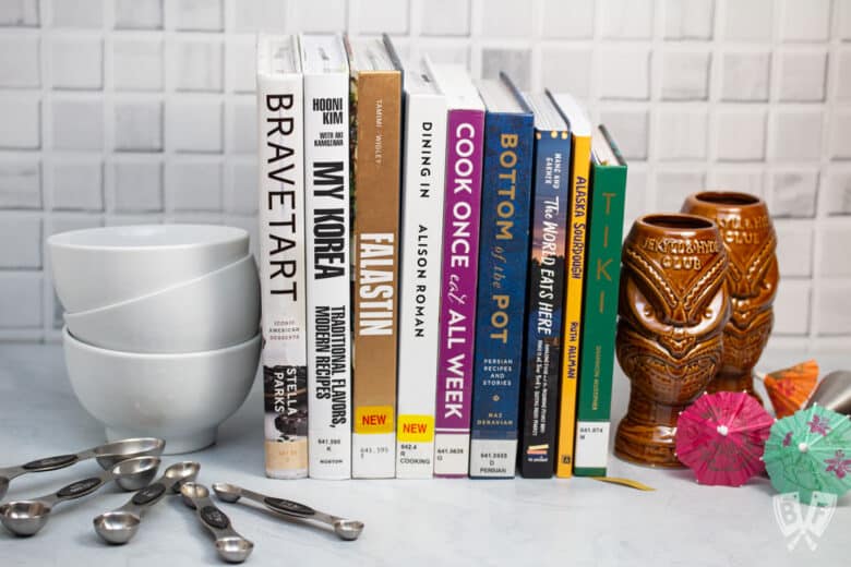 Assortment of cookbooks with measuring spoons, bowls, and tiki cocktail supplies alongside.