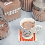 Homemade hot chocolate mix in jars surrounding a mug of hot cocoa with a spoon in it.