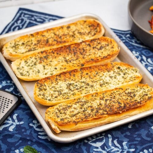 Overhead view of a baking sheet of golden brown garlic bread with plates of pasta and a chunk of Parmesan cheese alongside.
