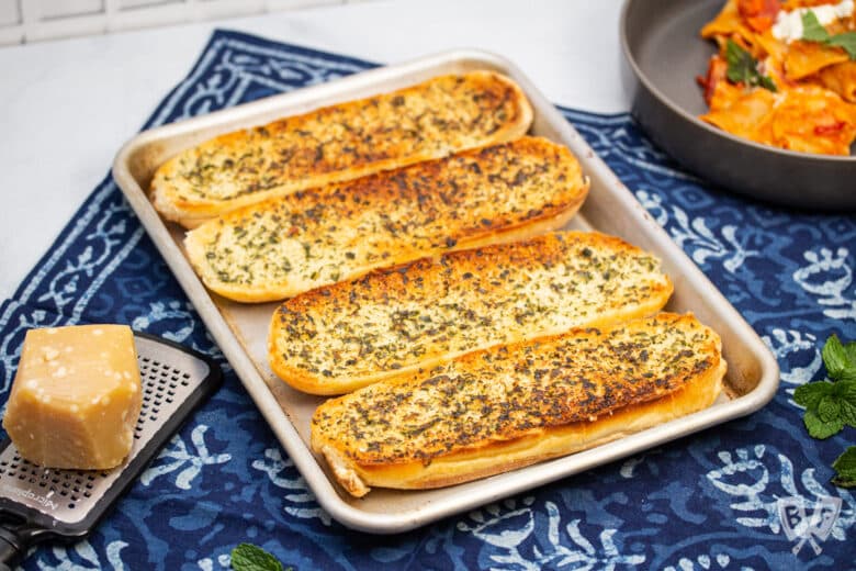 Overhead view of a baking sheet of golden brown garlic bread with plates of pasta and a chunk of Parmesan cheese alongside.