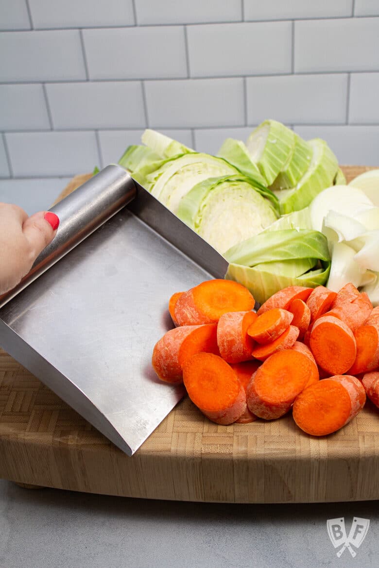 Scooping carrots from a cutting board onto a metal bench scraper with sides.