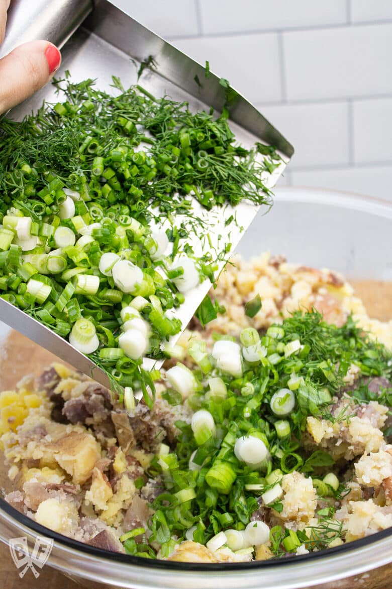 Adding fresh herbs and green onions to a bowl of smashed potatoes for herbed potato salad.