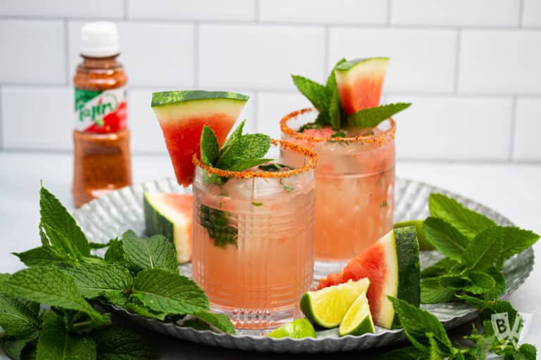 Tray of cocktails made with watermelon, mint, lime, and tequila with a bottle of Tajin seasoning in the background.