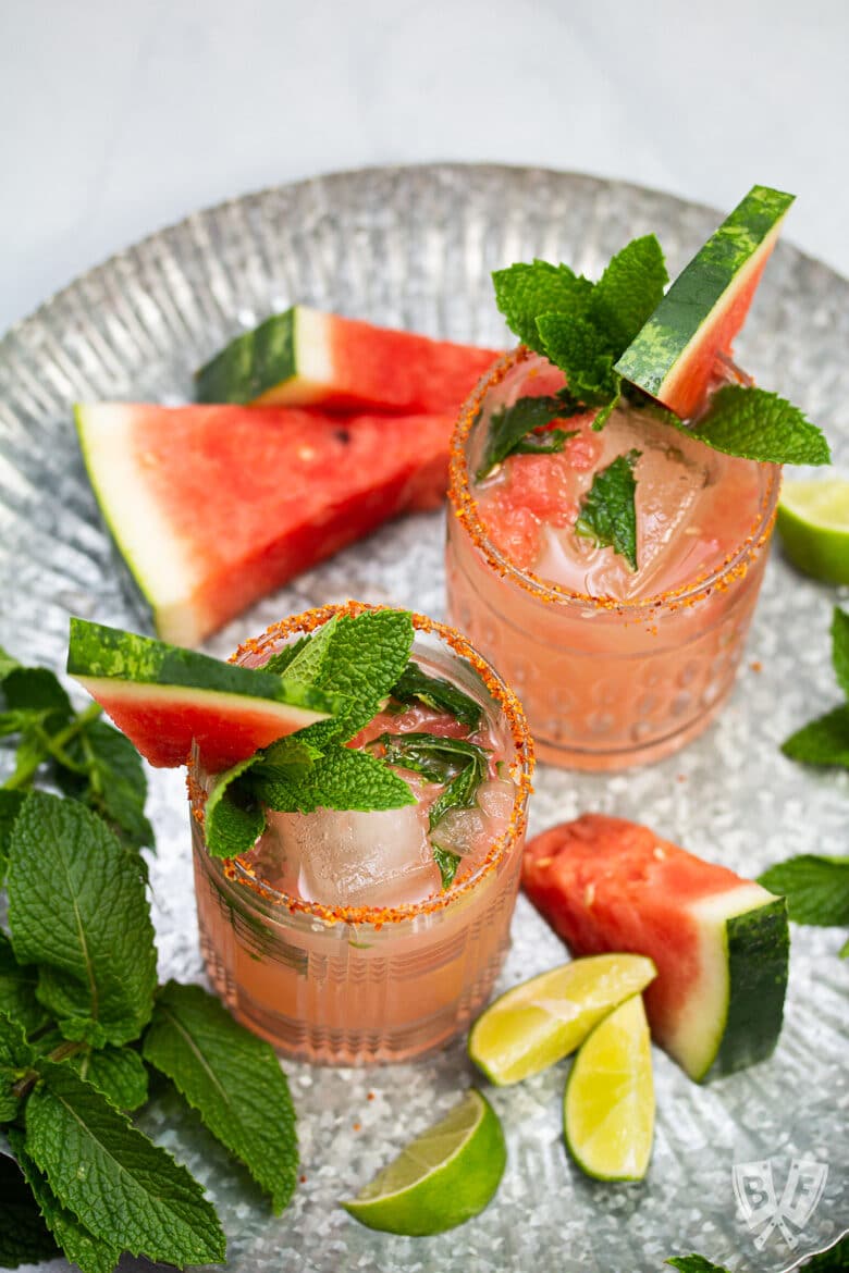 Watermelon cocktails on a tray surrounded by fresh watermelon, mint, and lime.