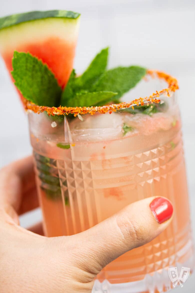 Hand holding a glass with a watermelon cocktail garnished with fresh mint and watermelon with a Tajin rim.