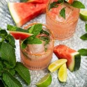 Watermelon cocktails on a tray surrounded by fresh watermelon, mint, and lime.