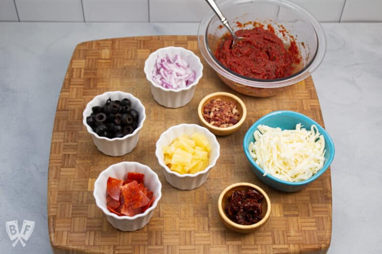 Board topped with bowls full of pizza sauce and toppings.