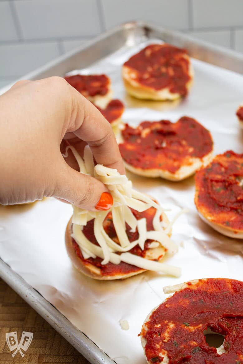 Adding cheese to pizza sauce topped mini bagels on a sheet pan.