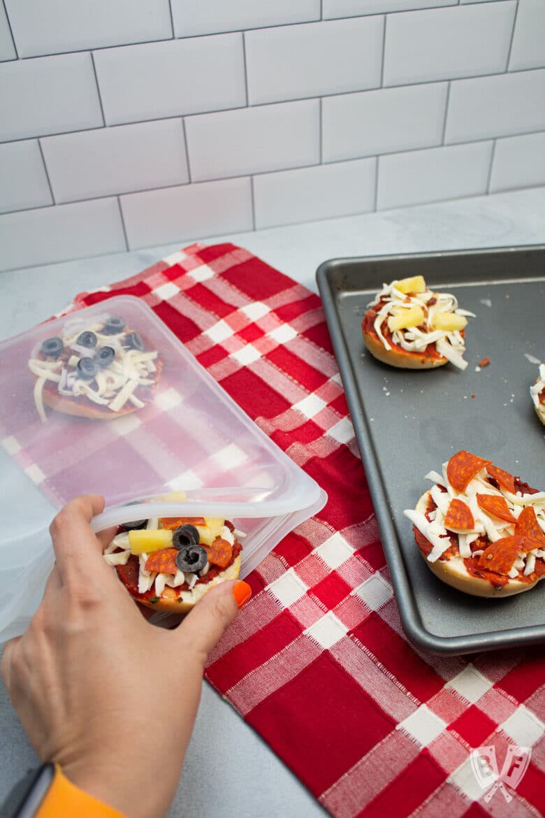 Tray of mini pizza bagels on the side with a hand placing some in a freezer bag.