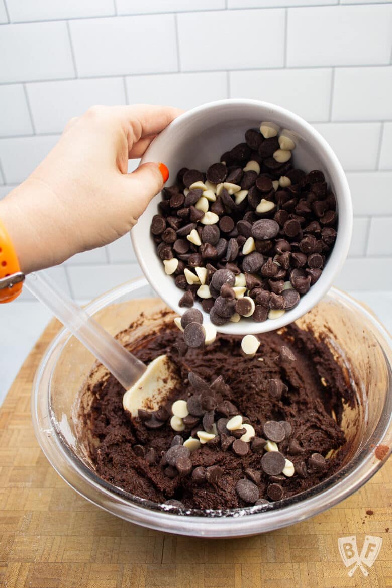 A variety of chocolate chips being poured into a bowl of chocolate cookie dough.