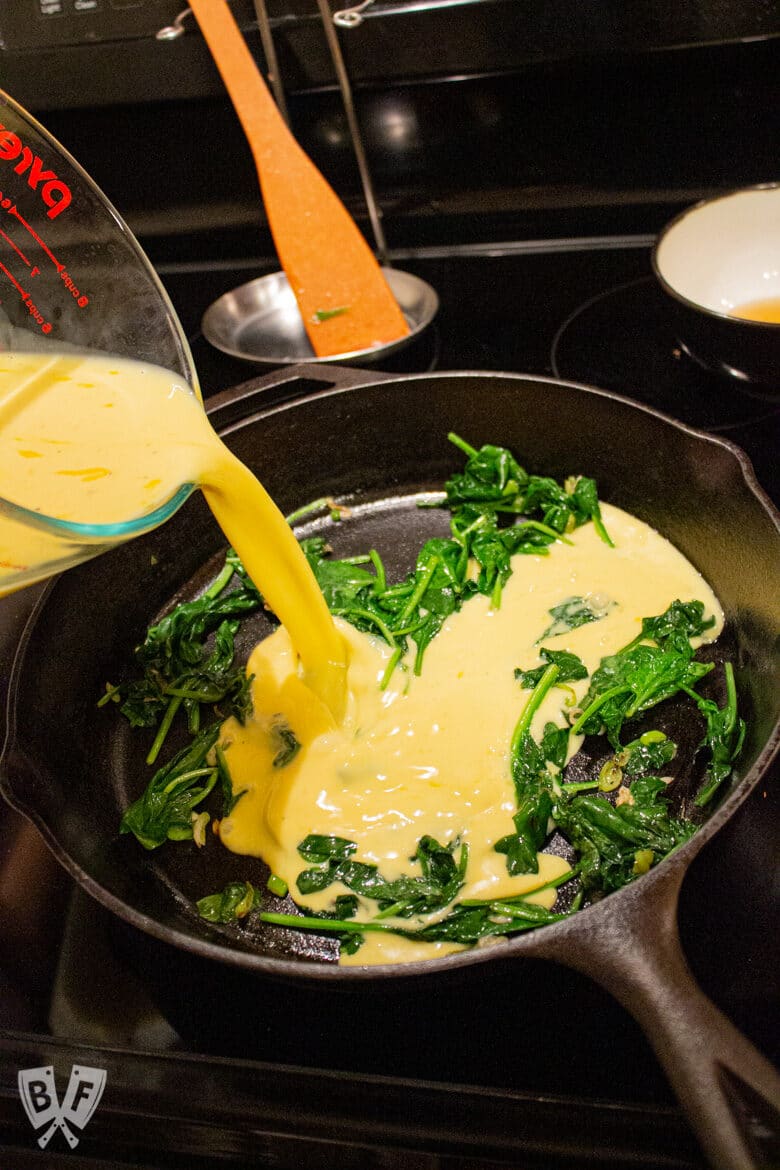 Pouring eggs and milk into a cast iron skillet with spinach.