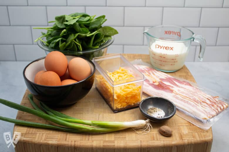 Spinach, eggs, green onion, cheddar cheese, bacon, milk, nutmeg, salt, and pepper on a butcher block.