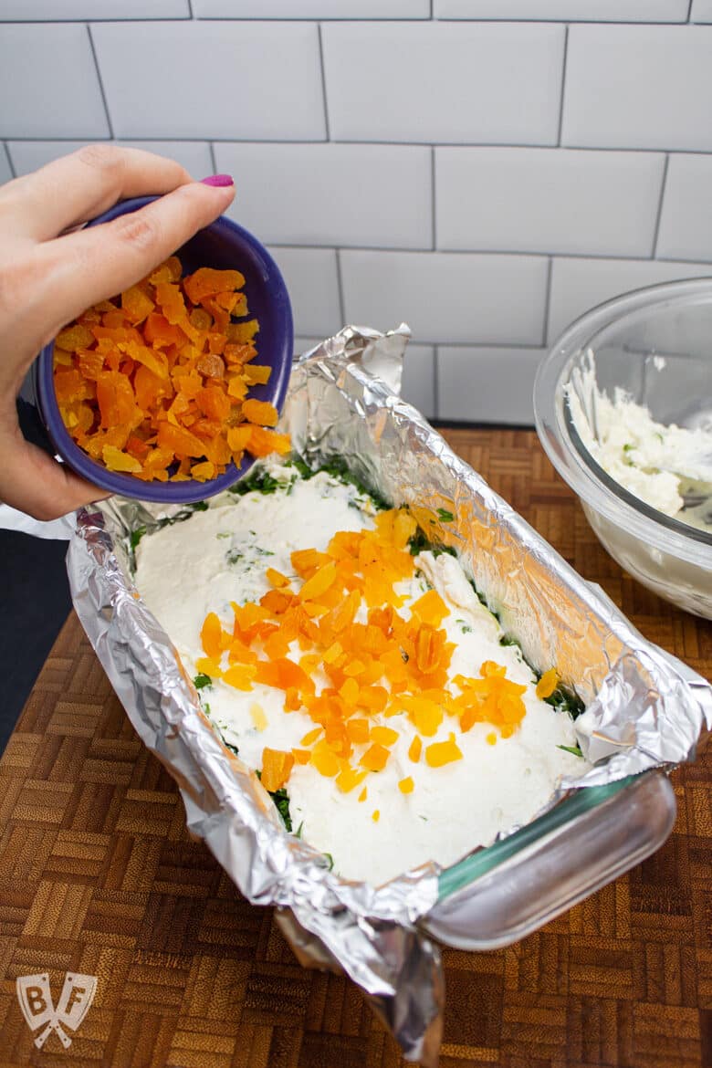 Sprinkling dried apricots onto a loaf pan full of layers of gorgonzola and cream cheese.