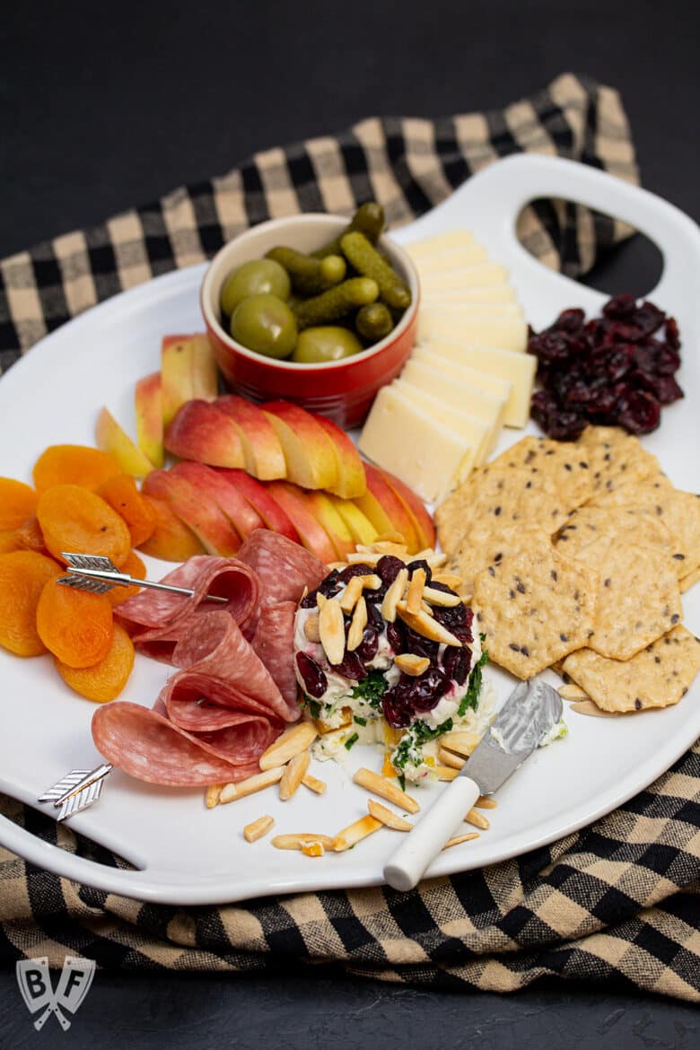Overhead view of a cheese board with assorted cheeses, meat, fruit, pickles, olives, and crackers.