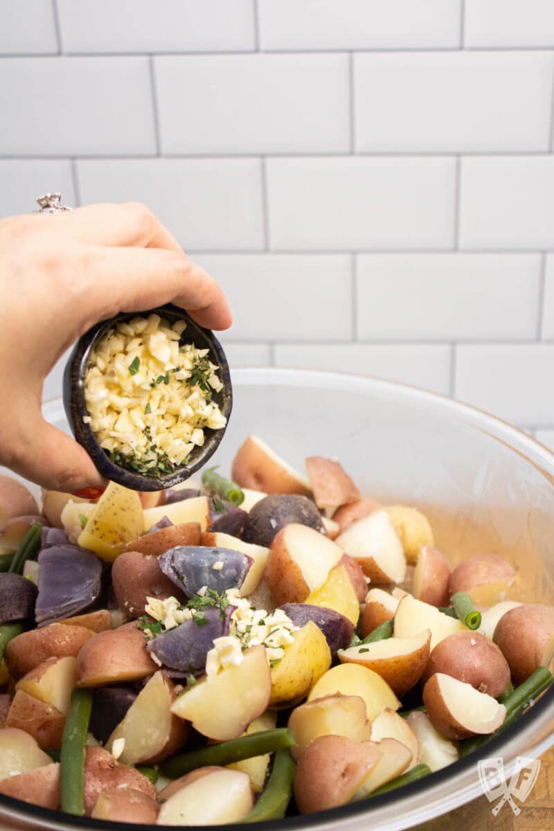 Pouring minced garlic and oregano into a bowl of potatoes and green beans.