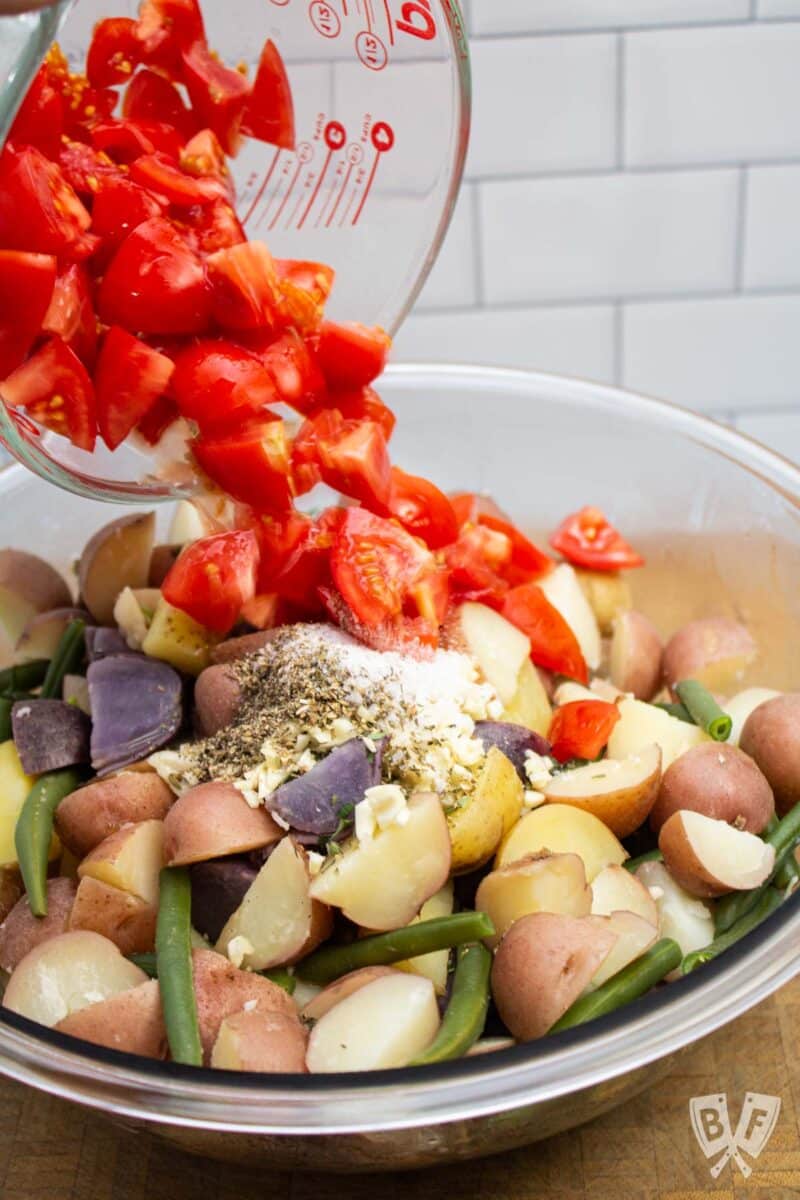 Pouring tomatoes into a bowl of potato salad with green beans.