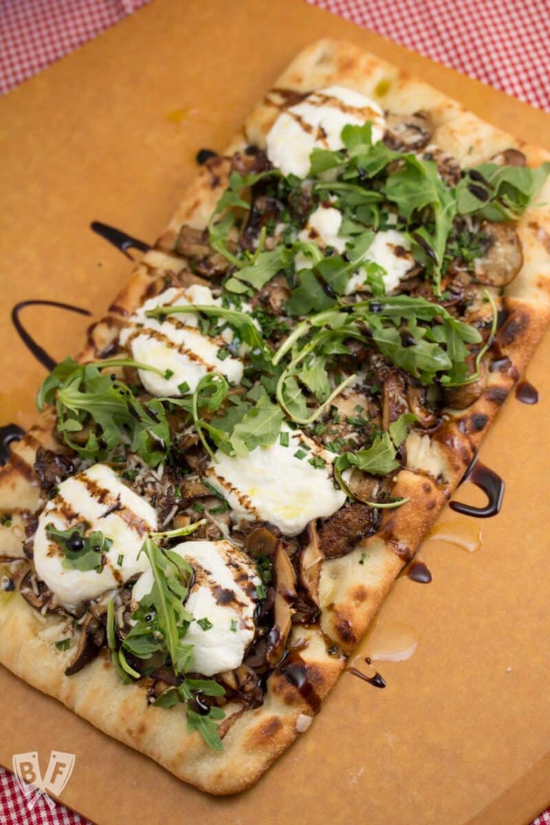 Flatbread on a cutting board with mushrooms, ricotta, baby arugula, and balsamic drizzle.