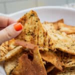 Holding a baked pita chip with a bowl full in the background.