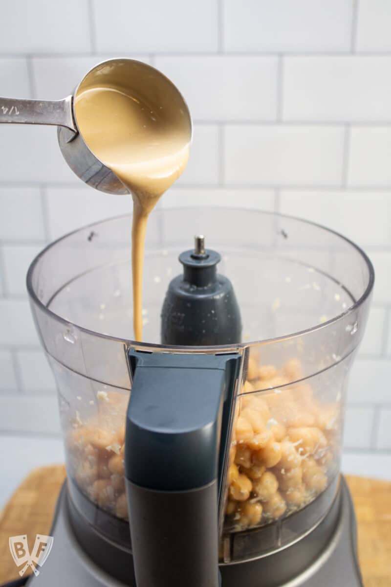 Adding tahini to hummus ingredients in a food processor.