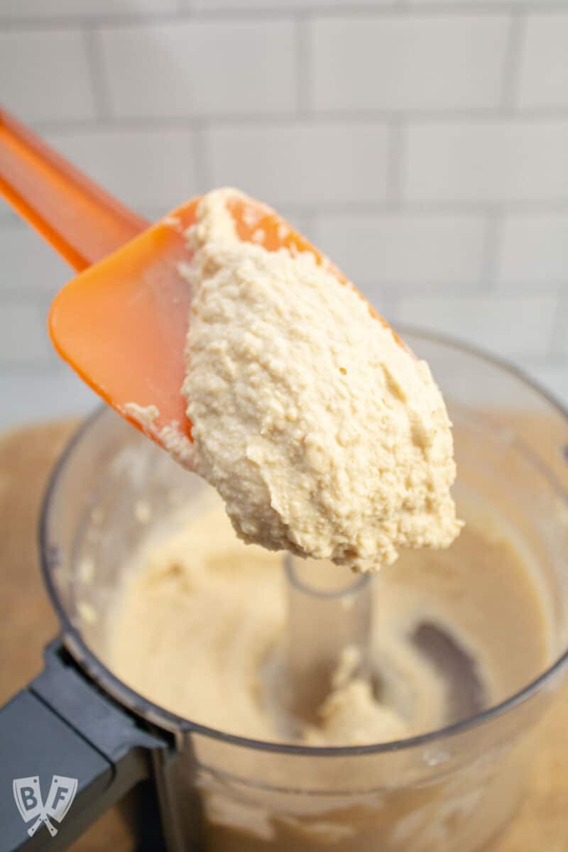 Scooping freshly made hummus from a food processor with a spatula.