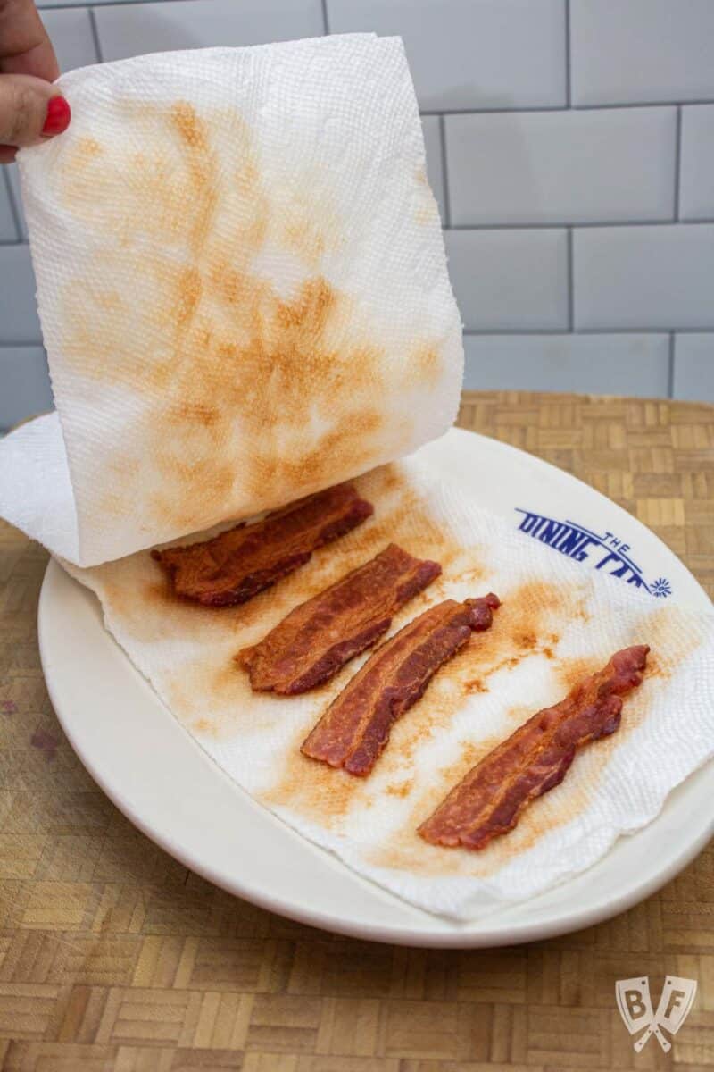 Lifting paper towels to reveal cooked bacon on a plate.