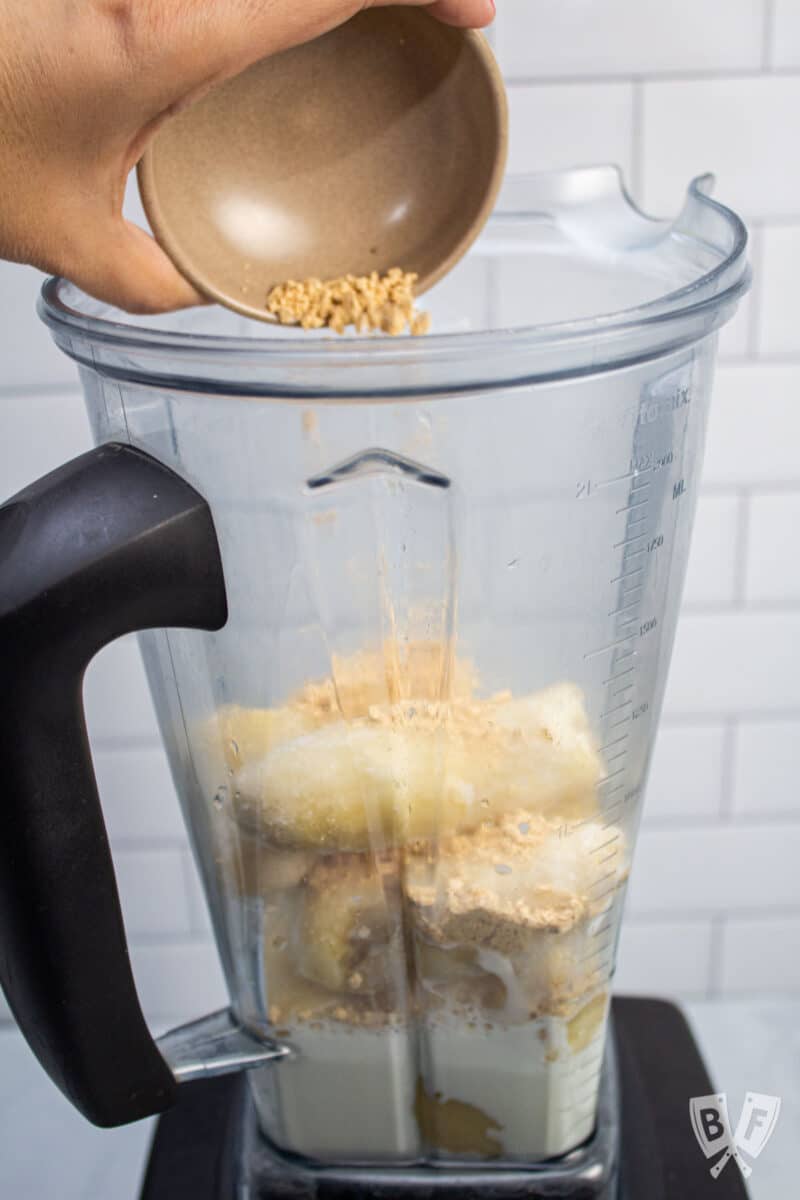 Adding peanut butter powder to a blender with frozen bananas and milk.