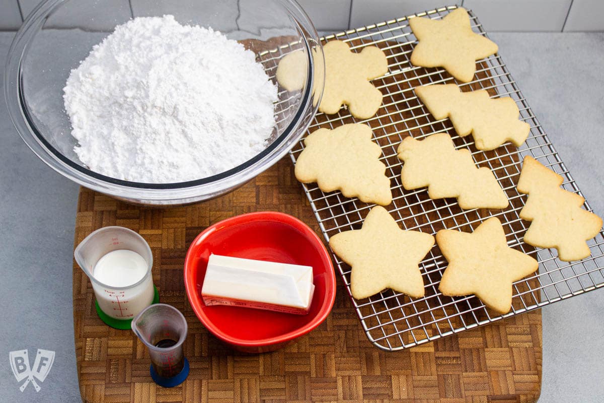 Icing ingredients on a cutting board with cut out sugar cookies on a cooling rack to the side.