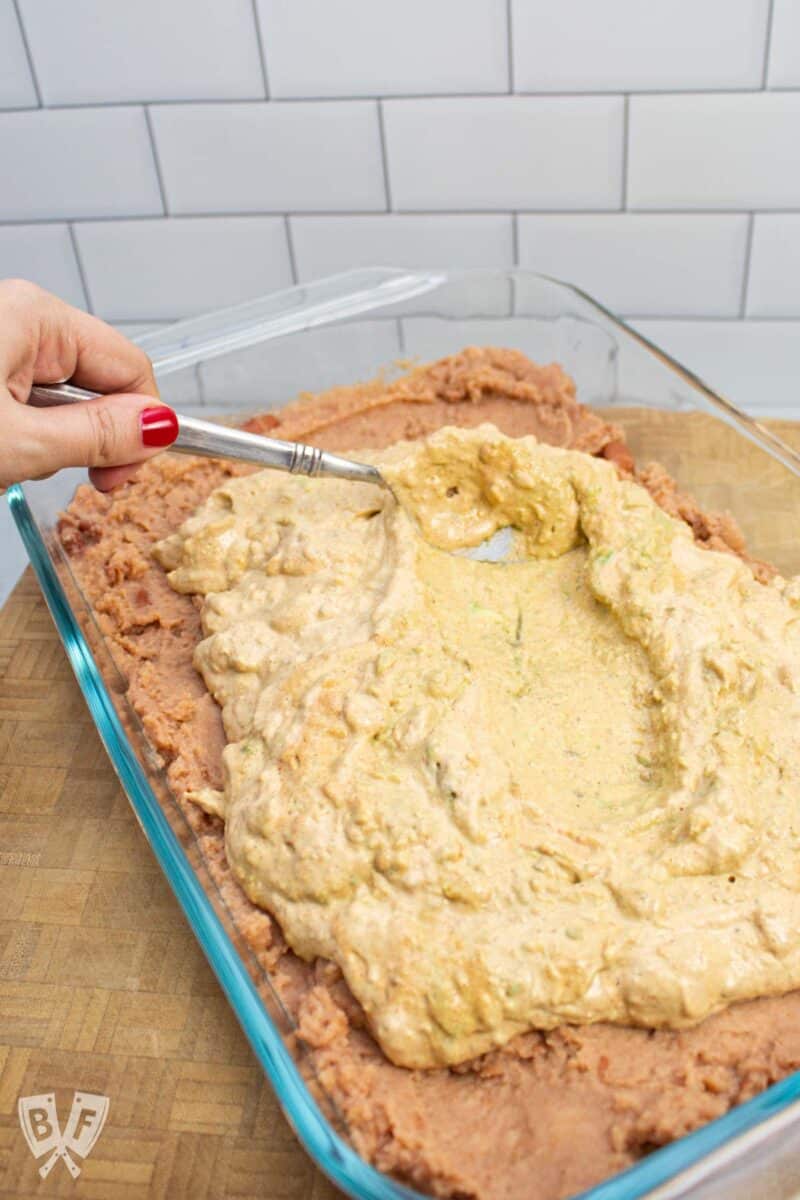 Smoothing a seasoned avocado mixture over refried beans in a serving dish for layered taco dip.