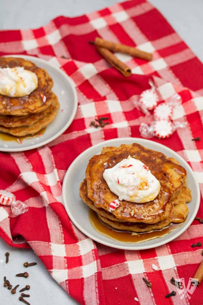 Two plates of gingerbread pancakes topped with peppermint whipped cream, crushed peppermint candies, and maple syrup.