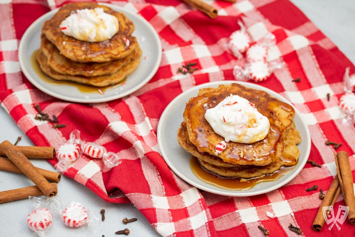 Two plates of gingerbread pancakes topped with peppermint whipped cream, crushed peppermint candies, and maple syrup.