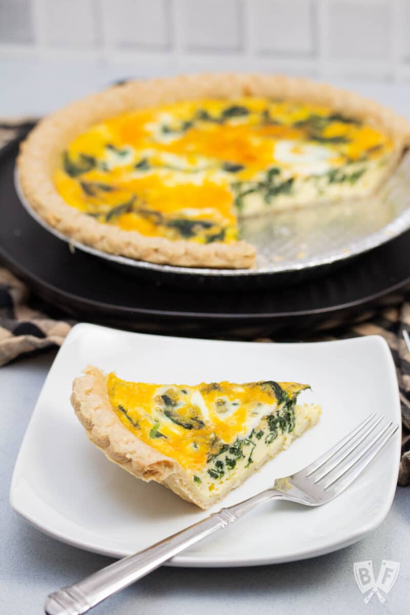 Slice of spinach and cheddar quiche on a plate next to the pie plate.