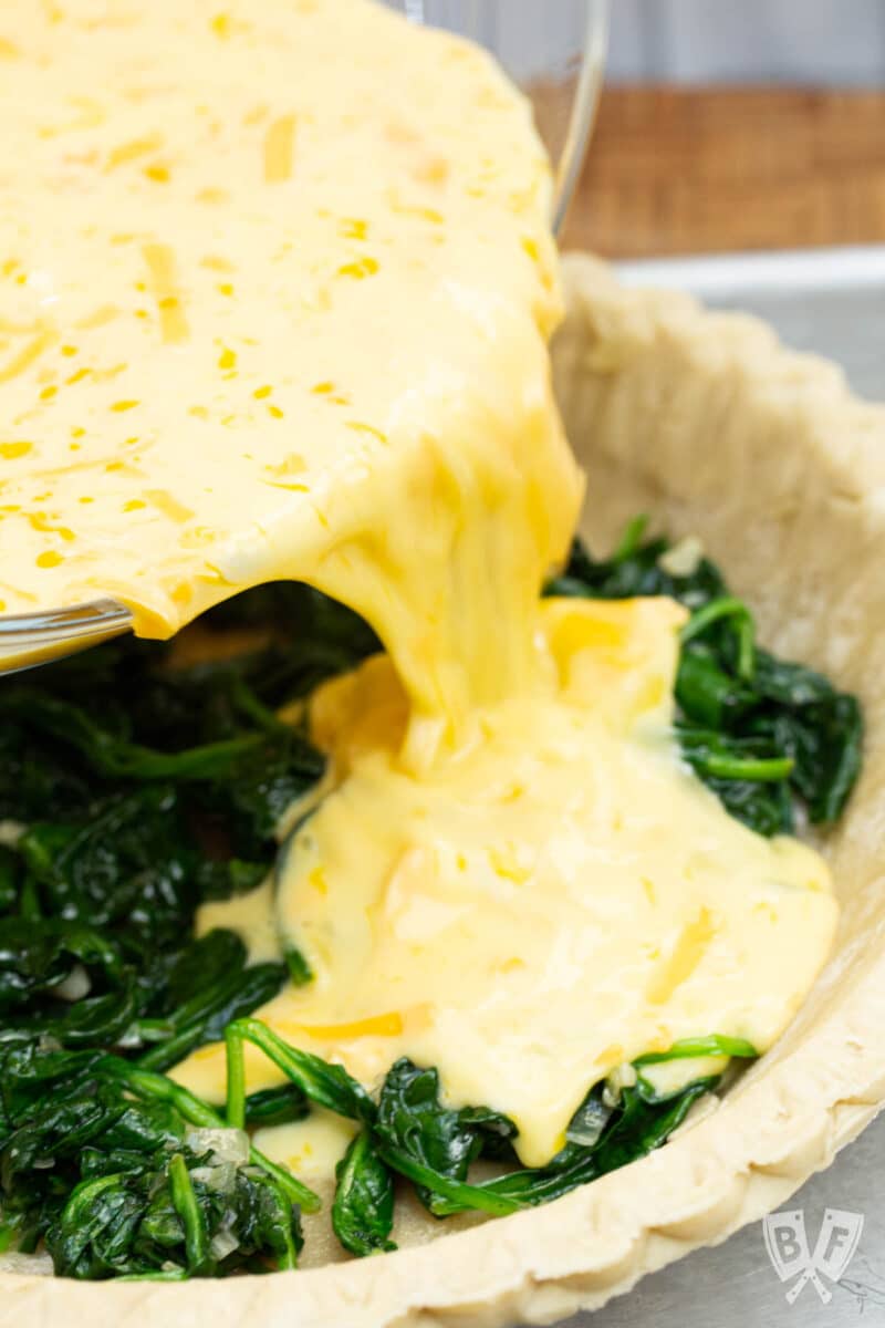 Pouring an egg mixture over sautéed spinach in a pie shell to make quiche.