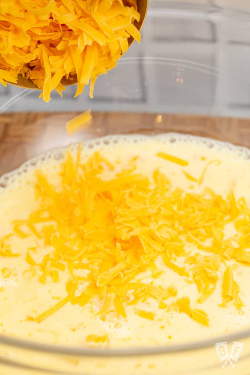 Pouring shredded cheddar into a bowl of eggs and milk for quiche.