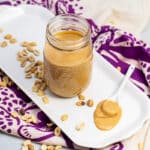 Platter with a jar of homemade peanut butter with peanuts scattered around and a spoon resting in a pile of peanut butter.