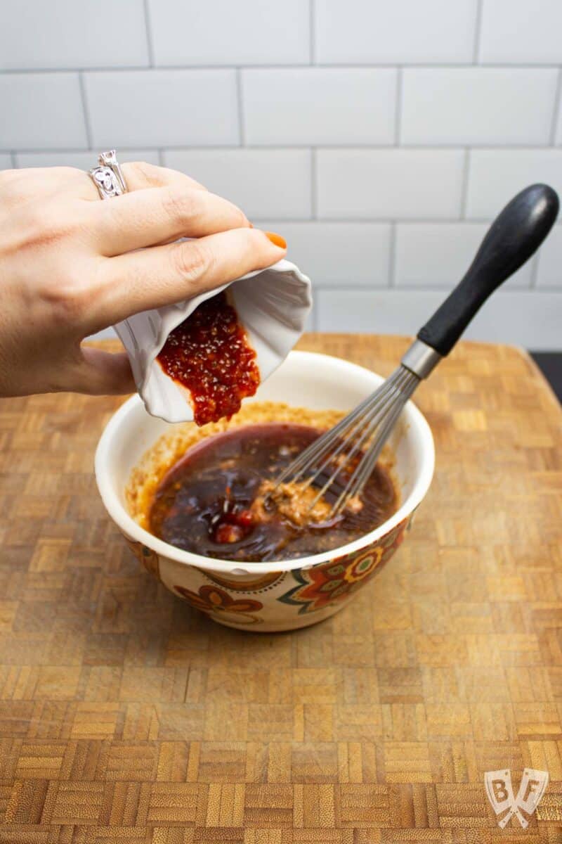 Adding chili garlic sauce to a bowl of peanut sauce ingredients.