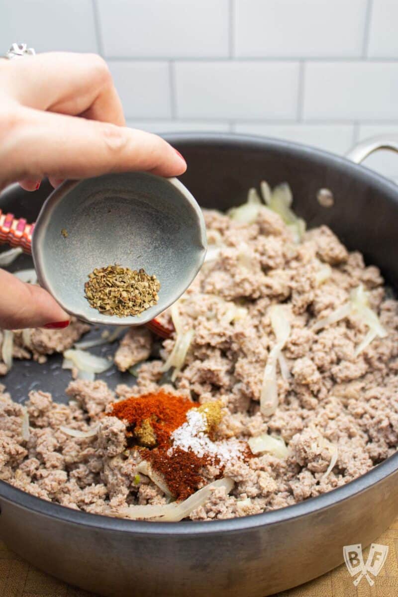 Adding dried oregano to a pan of turkey taquito filling.