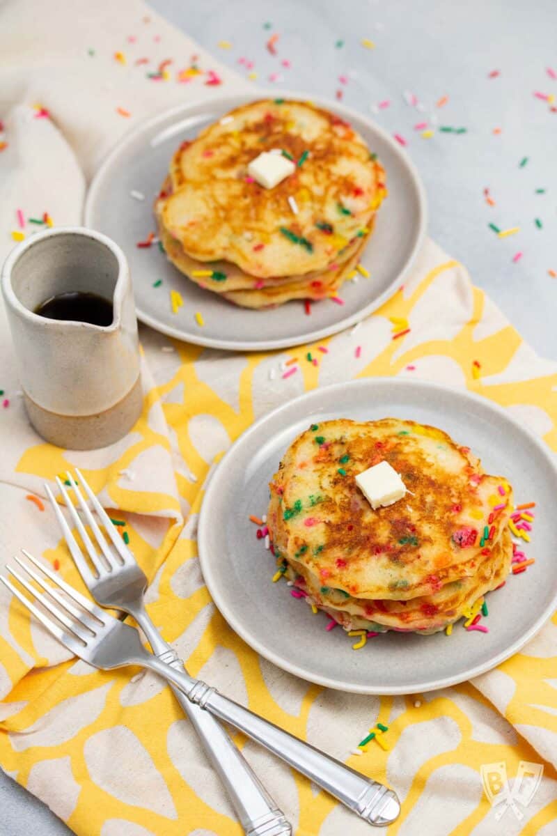 Plates of birthday cake pancakes with sprinkles served with maple syrup and butter.