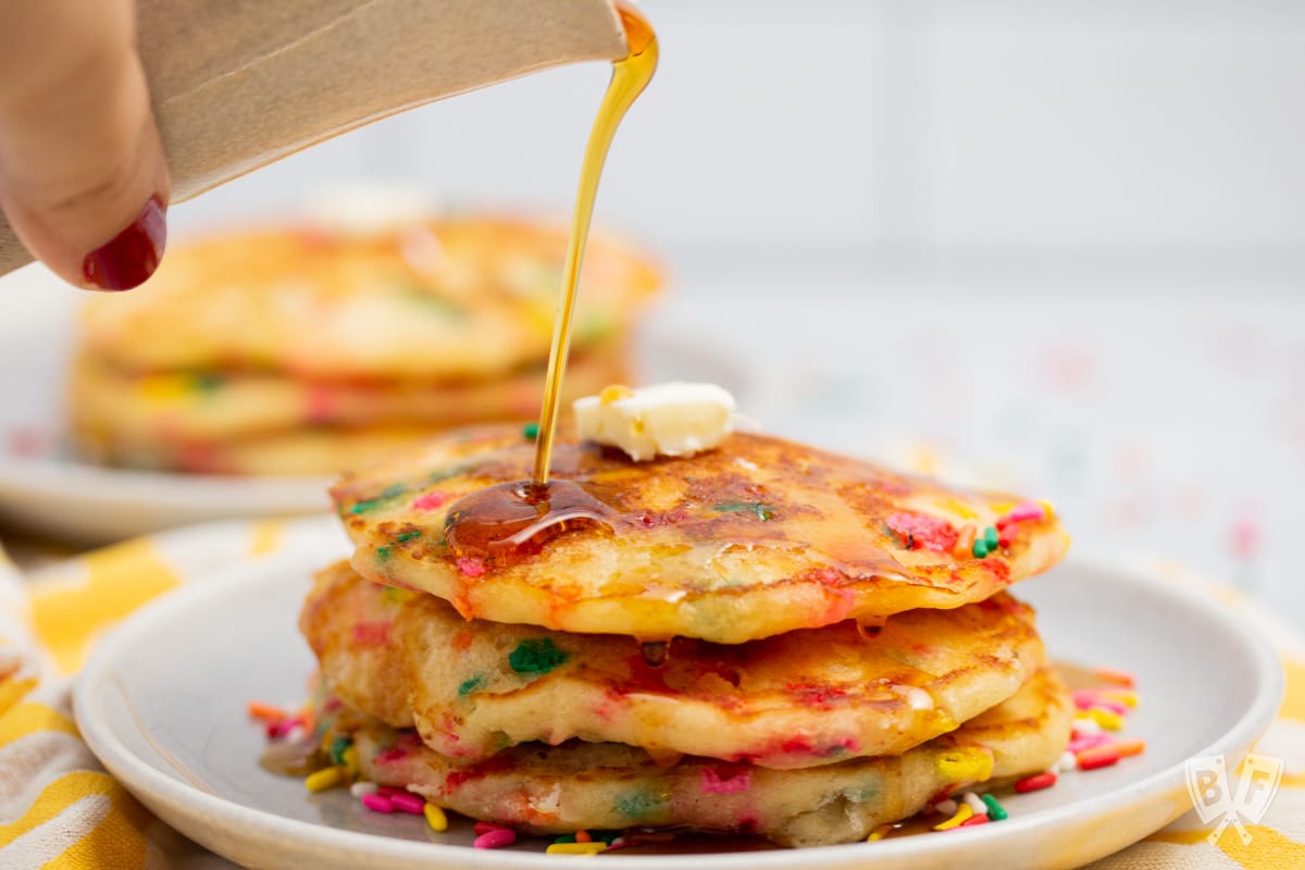 Close-up of drizzling maple syrup onto a stack of birthday cake pancakes.