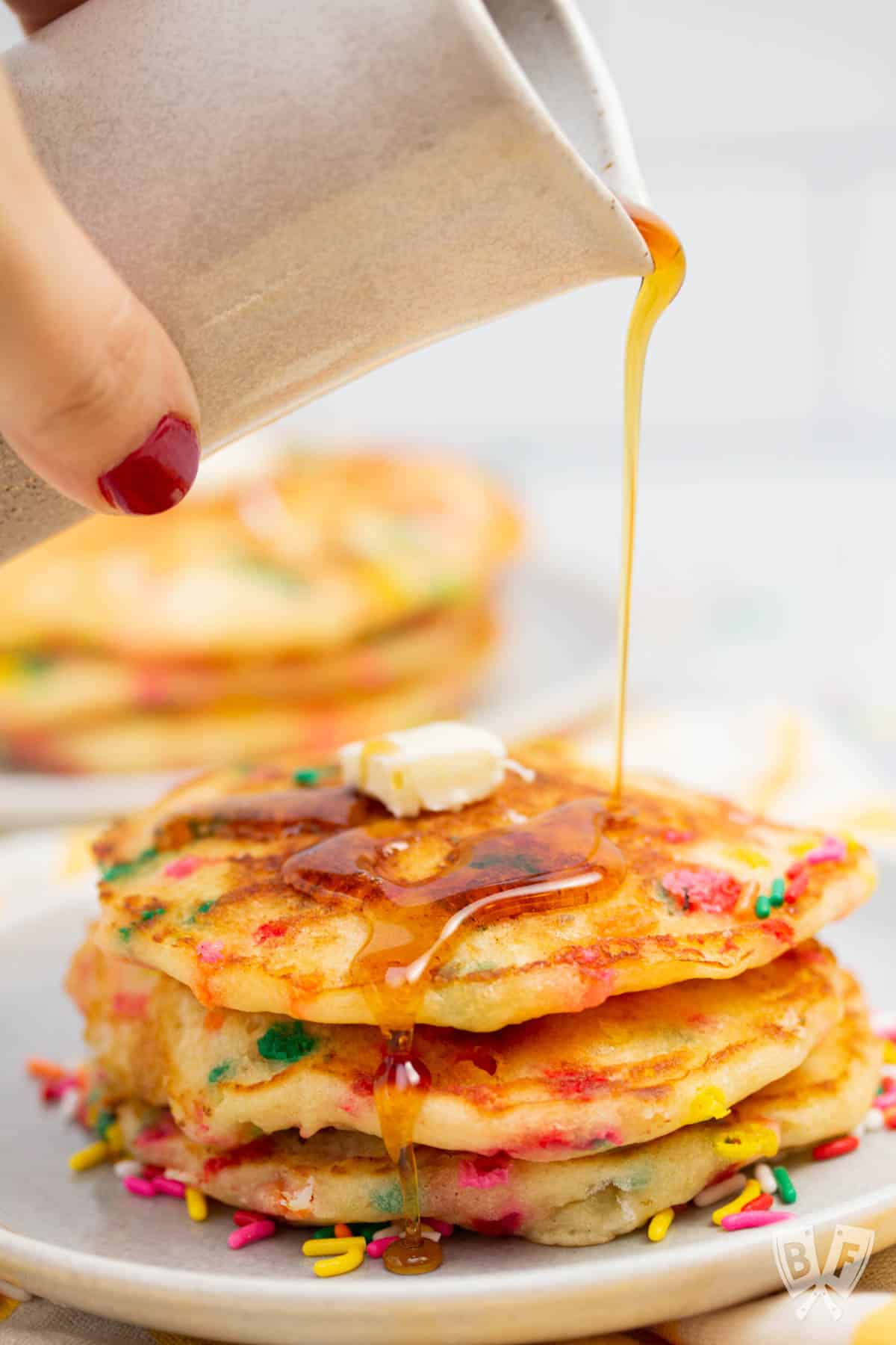 Drizzling maple syrup onto a stack of funfetti pancakes with sprinkles.