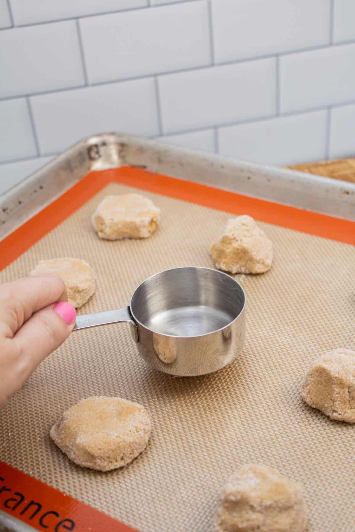 Flattening cookie dough balls on a baking sheet using an empty measuring cup.