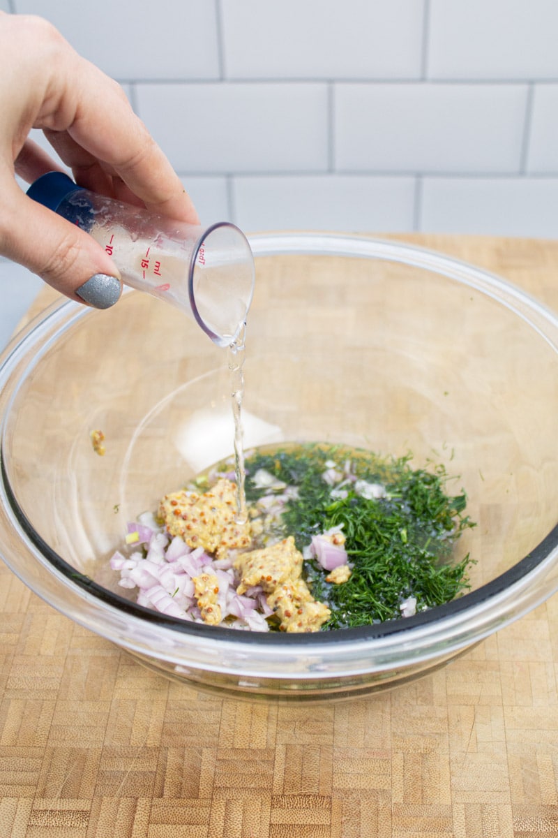 Pouring vinegar into a bowl with coarse ground mustard, dill, shallots, and olive oil.