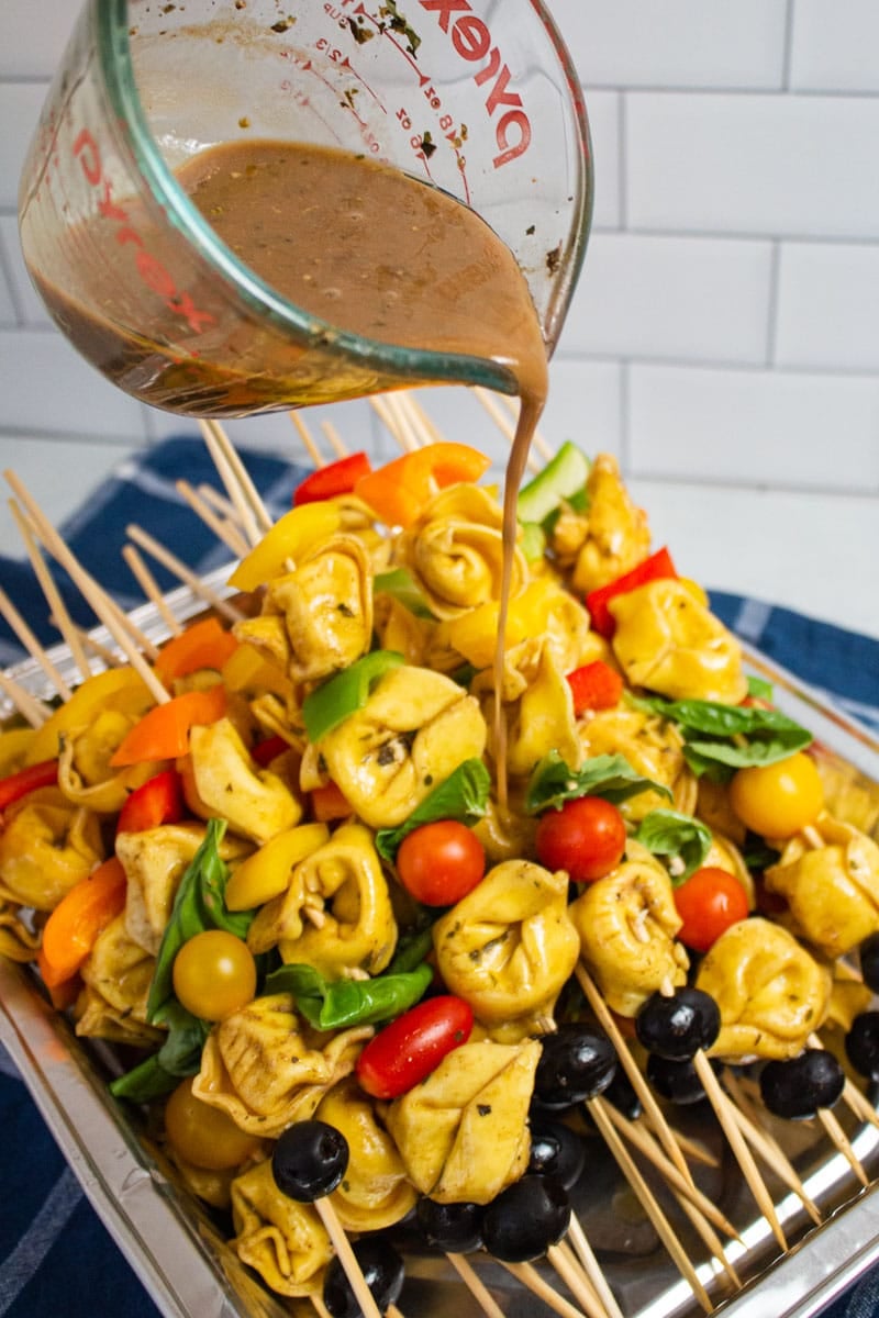 Drizzling creamy balsamic vinaigrette over a tray of pasta salad skewers.