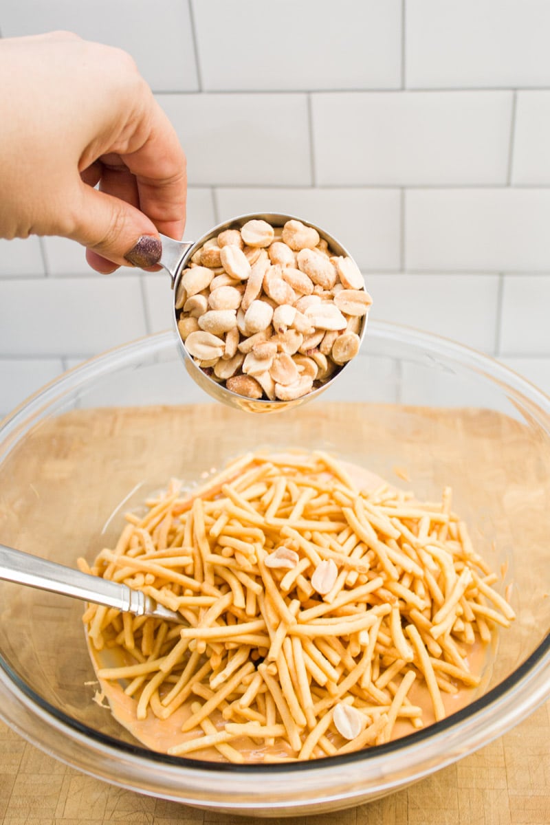 Adding peanuts to a bowl of chow mein noodles, melted butterscotch chips, and peanut butter.