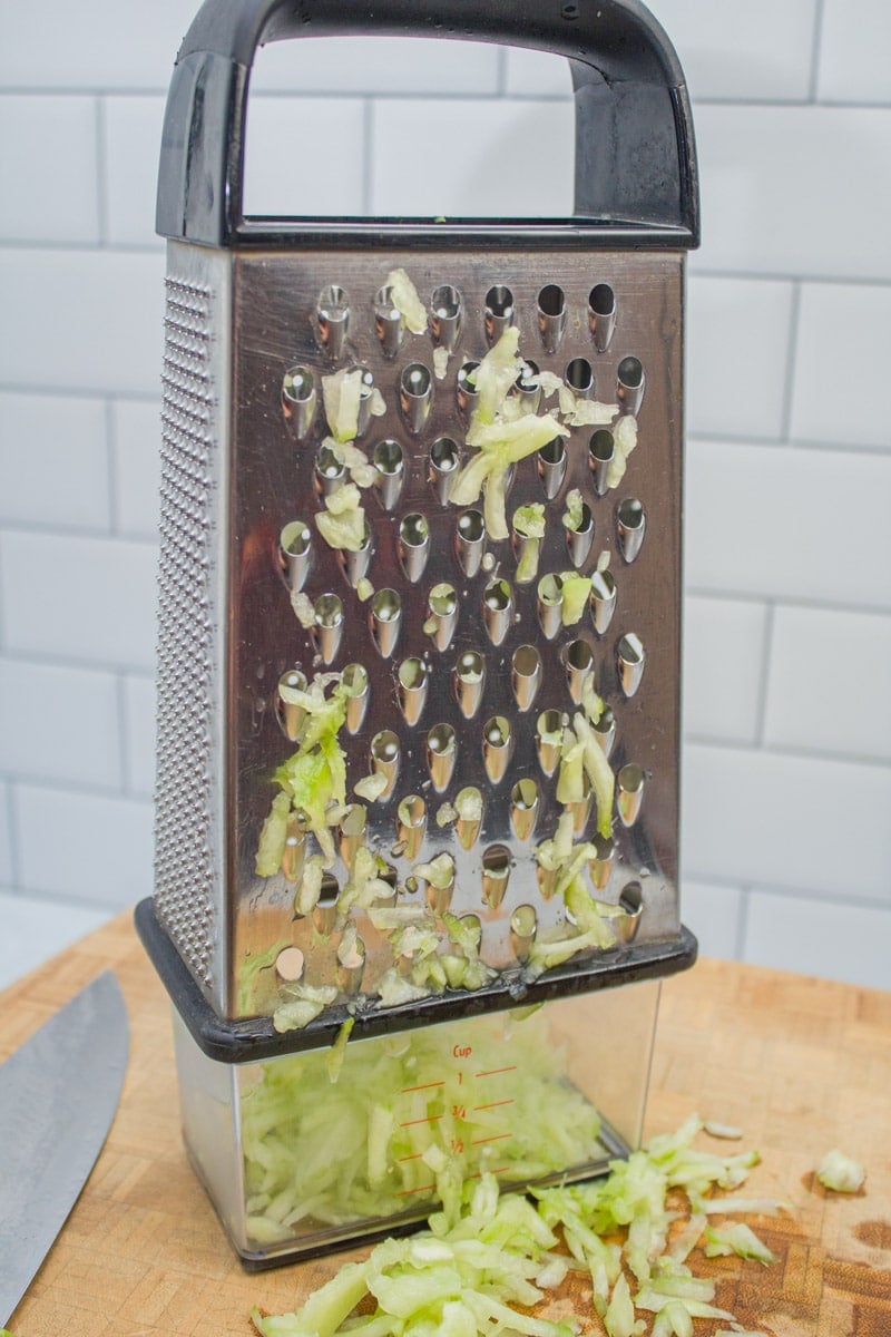 Box grater with shredded cucumber collected in a measuring cup.
